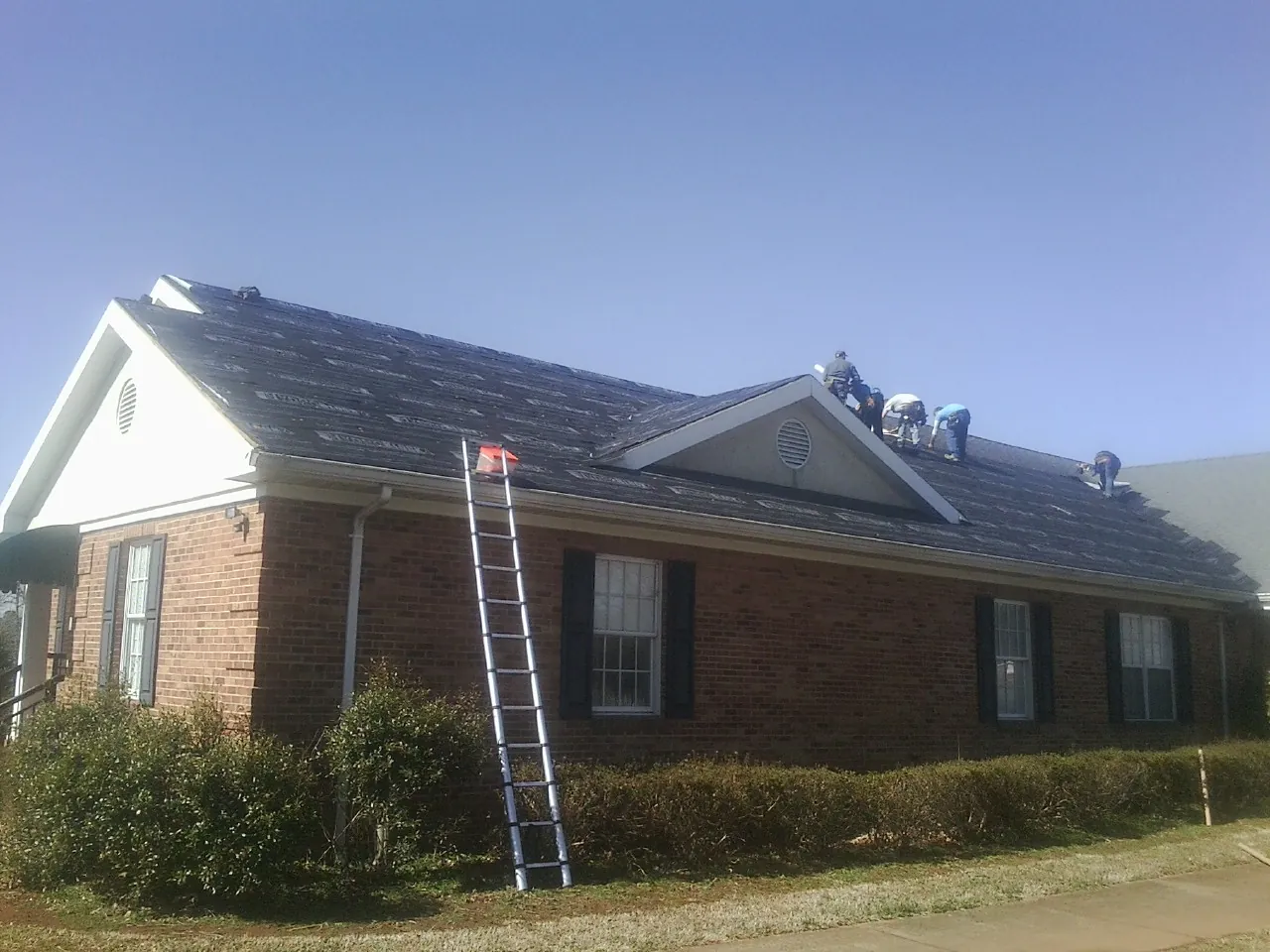 Crew tearing shingles off a brick ranch roof with a ladder set — Emergency Roof Repair work by our crew in Bondurant