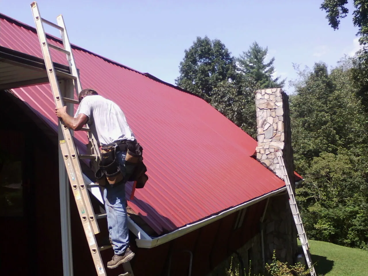 Roofer on a ladder beside a red metal roof and stone chimney — Soffit & Fascia Repair by Roofing Brothers in Bondurant